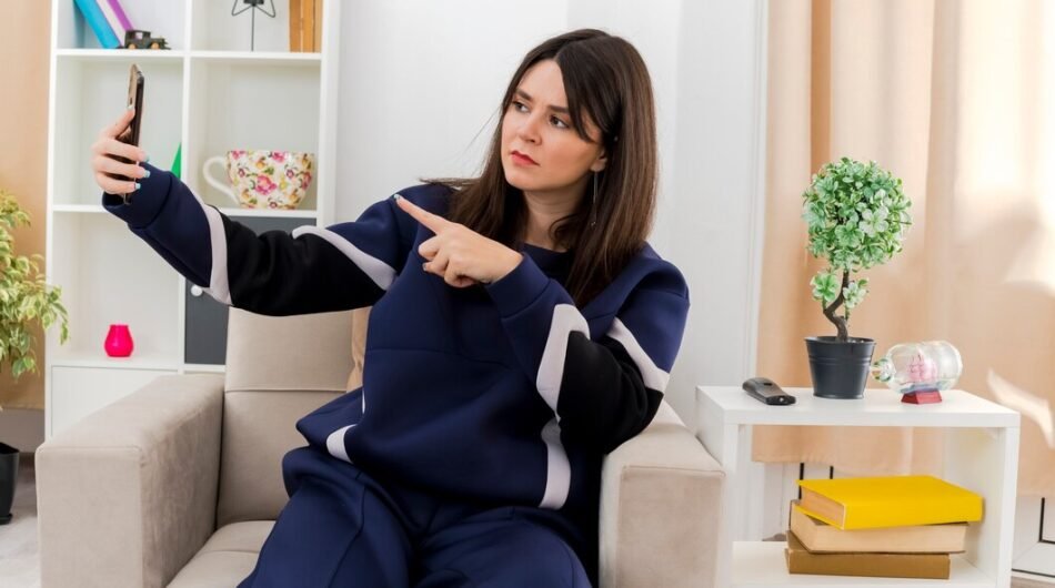 frowning-young-pretty-caucasian-woman-sitting-armchair-designed-living-room-pointing-phone-taking-selfie_141793-57847